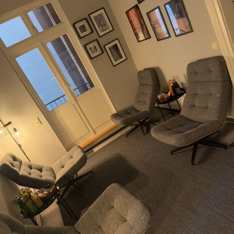 Alice Wonderline Tattoo Studio Gemütlicher Raum mit grauen Sesseln und Kunst an den Wänden, Fenster mit Aussicht.